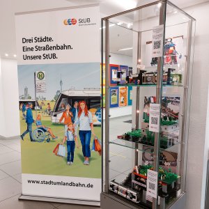 Modell der 6. Lego-Planungswerkstatt in einer Glasvitrine, daneben ein Roll Up zur Stadt-Umland-Bahn.